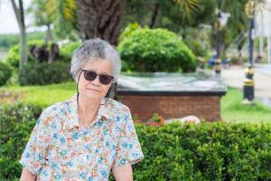 Portrait of a happy senior woman wearing sunglasses smiling and looking at the camera while sitting on a chair in the park. Space for text. Concept of aged people and relaxation photo