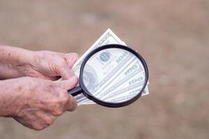 de cerca de mayor mujer manos participación dinero nosotros dolares y utilizar aumentador vaso en pie en jardín. selectivo enfocar. espacio para texto foto