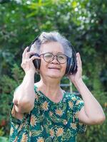 Senior woman with short gray hair wearing wireless headphones listening to a favorite song, smiling while standing in a garden. Concept of aged people and realxation photo