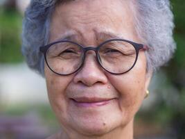 Close-up of face senior woman with short gray hair, wearing glasses, smiling and looking at the camera while standing in a garden. Concept of aged people and healthcare photo