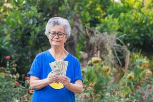 A senior woman with short gray hair, wearing glasses, smiling, holding US banknotes, and looking at the camera while standing in a garden. Concept of aged people and healthcare photo