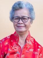 mayor mujer vistiendo un tradicional cheongsam qipao vestido, sonriente y mirando a el cámara mientras en pie en ligero naranja antecedentes. chino nuevo año concepto foto