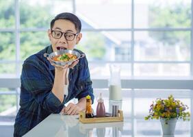 joven hombre vistiendo lentes sentado mirando a el frito huevo servido en un pan con vistoso coberturas servido en un cacerola. foto