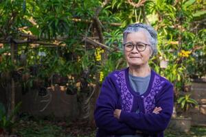 Portrait of a senior woman with short gray hair wearing glasses, smiling, arms crossed, and looking at the camera while standing outdoors. Space for text. Concept of aged people and healthcare photo