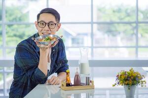 Young man wearing glasses holding the fried egg served on a pan with colorful toppings served on a pan while sitting on a chair. photo