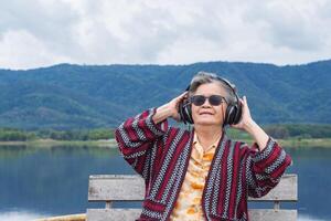 Portrait of a happy senior woman wearing wireless headphones listening to music for relaxing sitting at the side of the lake. Space for text. Concept of aged people and relaxation photo