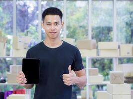 Young businessman wearing casual cloth holding a tablet, looking at the camera, and show fingers thumb up while standing in the office. Space for text. Concept of business and e-commerce photo