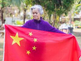 Portrait of an elderly Asian woman with short gray hair holding a Chinese flag while standing in a garden. Concept of aged people and relaxation photo