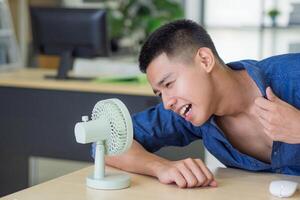 joven hombre utilizando un pequeño el plastico ventilador soplo a cara mientras sentado en el oficina. espacio para texto. concepto de clima foto