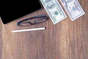 Top view of a stack of one hundred dollars, tablet, pencil and glasses on wooden desk. Space for text. Wood texture background. Business and finance concept photo