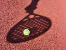 High angle of yellow ball placed on court and covered with shadow of crop person holding padel racket photo