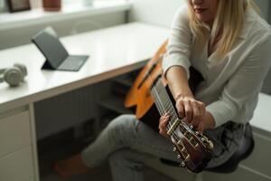sereno joven mujer en inalámbrico auriculares utilizando acústico musical instrumento mientras sentado con ordenador portátil foto
