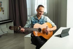 retrato de hombre tomando en línea guitarra lección mirando a ordenador portátil pantalla. retirado masculino aprendizaje a jugar guitarra acecho seminario web en computadora a hogar foto