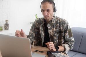 soldado en oficina. grave joven militar hombre mecanografía en ordenador portátil mientras trabajando en sede edificio. caucásico hombre en camuflaje uniforme es sentado en brillante habitación a mesa con cuaderno y gorra. foto