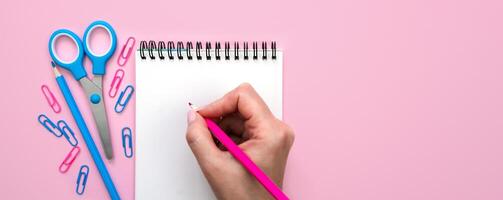 A woman's hand writes with a pink pencil in an empty notebook. Banner. Pink background. Flatlay composition. Top view. Copy space. photo