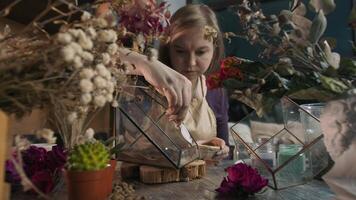 Talented woman surrounded by beautiful flowers in her studio creates a unique floral arrangement by covering the surface of a glass container with sand. This creative piece of art would fit perfectly in any interior. Medium shot. Slow motion. 4k video