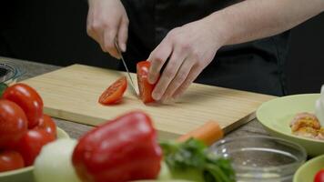 Process of preparing dinner. A man confidently cuts a juicy and ripe tomato into slices on a cutting board, surrounded by other vegetables for a salad. Cooking master class. Medium shot. Slow motion. 4k. video