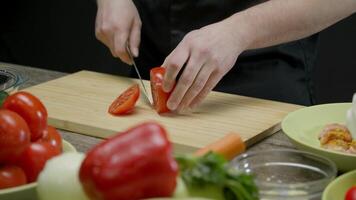 processi di preparazione cena. un' uomo con fiducia tagli un' succoso e maturo pomodoro in fette su un' taglio asse, circondato di altro verdure per un' insalata. cucinando maestro classe. medio sparo. lento movimento. 4k. video