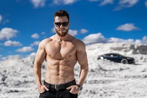 Brutal man with perfect body and strong abs wears sunglasses and looks to the camera. White quarry and luxury black car background. photo