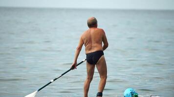 hombre cenar mar. mayor hombre aprende a paleta cenar en pie en tablero en abierto mar Oceano en soleado día. verano fiesta vacaciones y viaje concepto. espalda vista. lento movimiento video