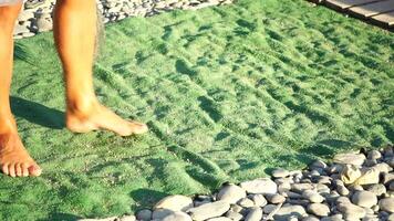 irreconocible hombre barriendo el verde alfombra de artificial césped en el playa desde guijarros con escoba. video