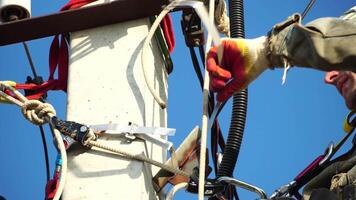 Skilled electrician in helmet fixes wires standing on ladder near high pole against blue sky on summer day backside view. Electrical service and mounting on the pole. Slow motion video