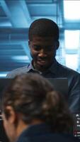 Vertical Portrait of cheerful african american manager supervising team in server room providing computing resources for workloads. Jolly supervisor oversees technicians in data center mending supercomputers video