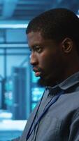 Vertical African american IT expert in server farm ensuring valuable data remains shielded from threats. Man safeguarding data center hardware against unauthorized access and vulnerabilities, securing system video