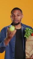 verticale verticale sur de soi homme jonglerie avec un éco amical vert Pomme sur caméra, en présentant mûr localement grandi des fruits. Jeune adulte en jouant autour et porter papier sac avec biologique produire. caméra b. video