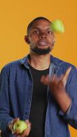 Vertical Playful person juggling in the studio with ripe green apples, keeping it in the air with continuous motion by throwing and catching. Confident skilled guy having fun with organic fruits. Camera B. video