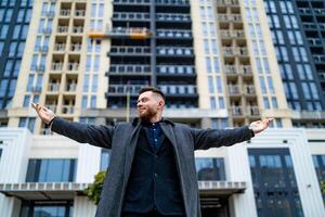 Portrait of a charming young businessman dressed in suit and coat standing outside a modern building with widely opened arms. photo