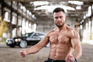 músculo sexy desnudo joven hombre sin camisa poses en abandonado almacén con negro coche en el borroso antecedentes. concepto foto. foto