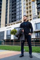 Man holds coat in hand and poses to the camera in ellegant dark suit. Fasion businessman with modern business center in the background. photo