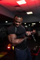 Sportsman in black sport clothes doing workout for hands and lifts weight on sport equipment. Muscular african american. photo