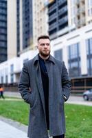 Fashionable man in coat stands near office centre and watching forward. Business person keeps calm and thinking. Successful concept. Thoughtful and resolute look of handsome man in big town. photo