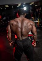High and shirtless man poses with very muscular back to the camera. African american bodybuilder with perfect body. Dark gym background. photo