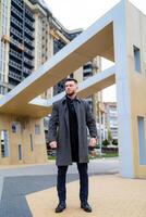 calle negocio estilo. hombre en un elegante oscuro traje y gris abrigo. hermoso joven caucásico hombre es en pie en un central calle con arriba a fecha arquitectura. foto