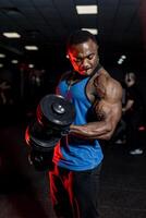 profesional africano americano deportista posando en cámara en deporte ropa y pesas en mano. negro gimnasio antecedentes. muscular Perfecto cuerpo. foto