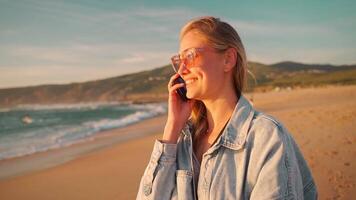 heiter Frau reden auf Smartphone während genießen auf Strand während Sonnenuntergang video