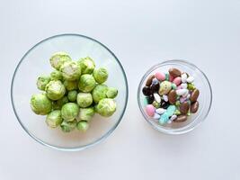 Fresh Brussels sprouts in glass bowl next to bowl of colorful candy on white background. Healthy versus indulgent food concept. photo