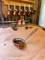 Copper cezve with fresh Turkish coffee on sandy stove, traditional copper coffee pots on wooden rack in background, symbolizing the authenticity of Middle Eastern coffee traditions. photo