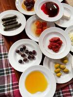 Assorted Turkish breakfast dishes with olive oil, honey, jams, rolled grape leaves, olives, feta cheese, and tomato sauce on white plates, traditional patterned tablecloth. Top view. photo