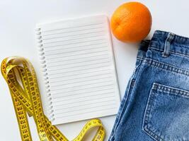 sano estilo de vida y dieta planificación concepto con naranja, medición cinta, blanco computadora portátil, y mezclilla pantalones en blanco antecedentes con Copiar espacio. parte superior vista. foto