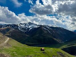 Solitary house in verdant valley with snow capped mountains under dynamic sky with billowing clouds, depicting isolation and beauty of nature. photo