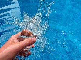 Hand holding clear glass bottle partially filled with water against sparkling blue water background with empty space for text. Clean water concept. photo