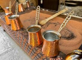 Traditional Turkish coffee in copper turks with long handles on tray with sand. Real coffee preparation, close up with selective focus. photo