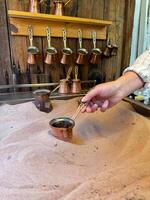 Preparing Turkish coffee in traditional copper pot on sandy stove, with hand holding the handle, copper cezves and wooden wall rack in the background. Turkish coffee culture concept. photo