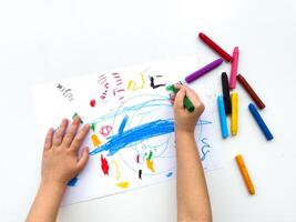 Childs hands drawing with colorful wax crayons on white paper, top view. Creative art concept for educational and developmental activities. photo