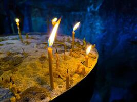 iluminado velas en arena lleno vela poseedor en Iglesia en contra oscuro pared. religioso o espiritual ceremonia con parpadeando llamas y calentar luz de una vela. ardiente velas en iglesia. foto