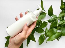 Close up of middle age woman hand holding white lotion bottle with green leaves on white background. Skincare and natural beauty concept. Healthcare, wellness, and organic product poster. photo